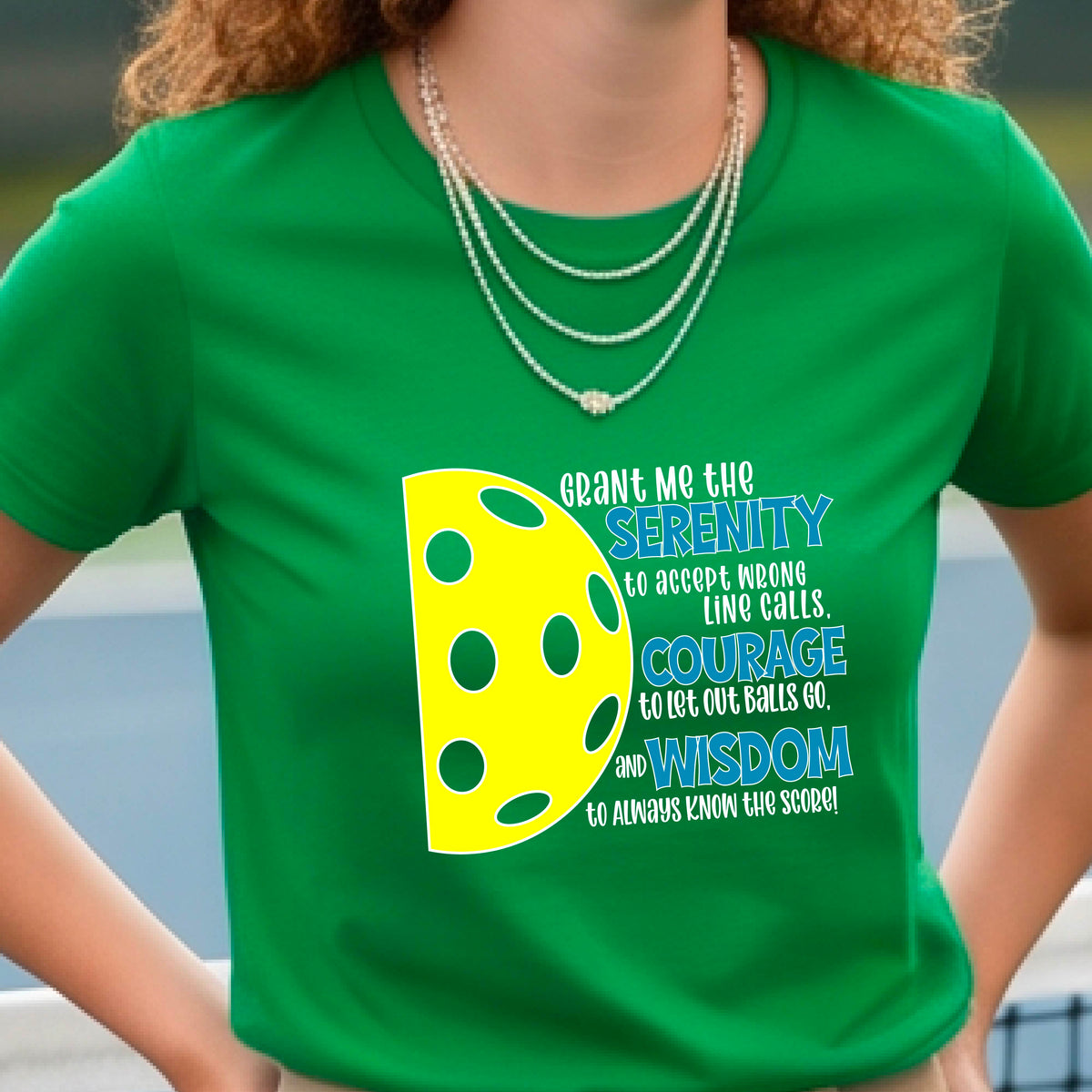 A woman on a pickleball court wearing a green short-sleeve t-shirt with a graphic of the Serenity Prayer made for pickleball:  “Grant me the serenity to Accept Wrong Line Calls; Courage to Let Out Balls Go; and, Wisdom to Always Know the Score” next to a half-pickleball icon, standing on a pickleball court.