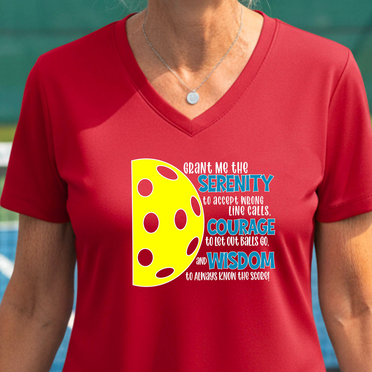 A woman on a pickleball court wearing a red short-sleeve v-neck t-shirt with a graphic of the Serenity Prayer made for pickleball:  “Grant me the serenity to Accept Wrong Line Calls; Courage to Let Out Balls Go; and, Wisdom to Always Know the Score” next to a half-pickleball icon, standing on a pickleball court.