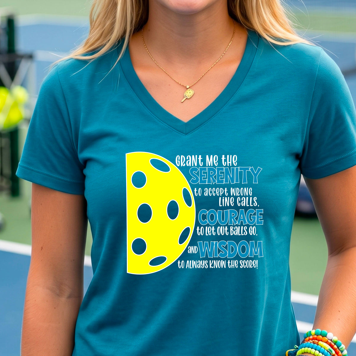 A woman on a pickleball court wearing a teal short-sleeve v-neck t-shirt with a graphic of the Serenity Prayer made for pickleball:  “Grant me the serenity to Accept Wrong Line Calls; Courage to Let Out Balls Go; and, Wisdom to Always Know the Score” next to a half-pickleball icon, standing on a pickleball court.