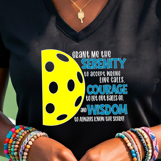 A woman on a pickleball court wearing a black short-sleeve v-neck t-shirt with a graphic of the Serenity Prayer made for pickleball:  “Grant me the serenity to Accept Wrong Line Calls; Courage to Let Out Balls Go; and, Wisdom to Always Know the Score” next to a half-pickleball icon, standing on a pickleball court.