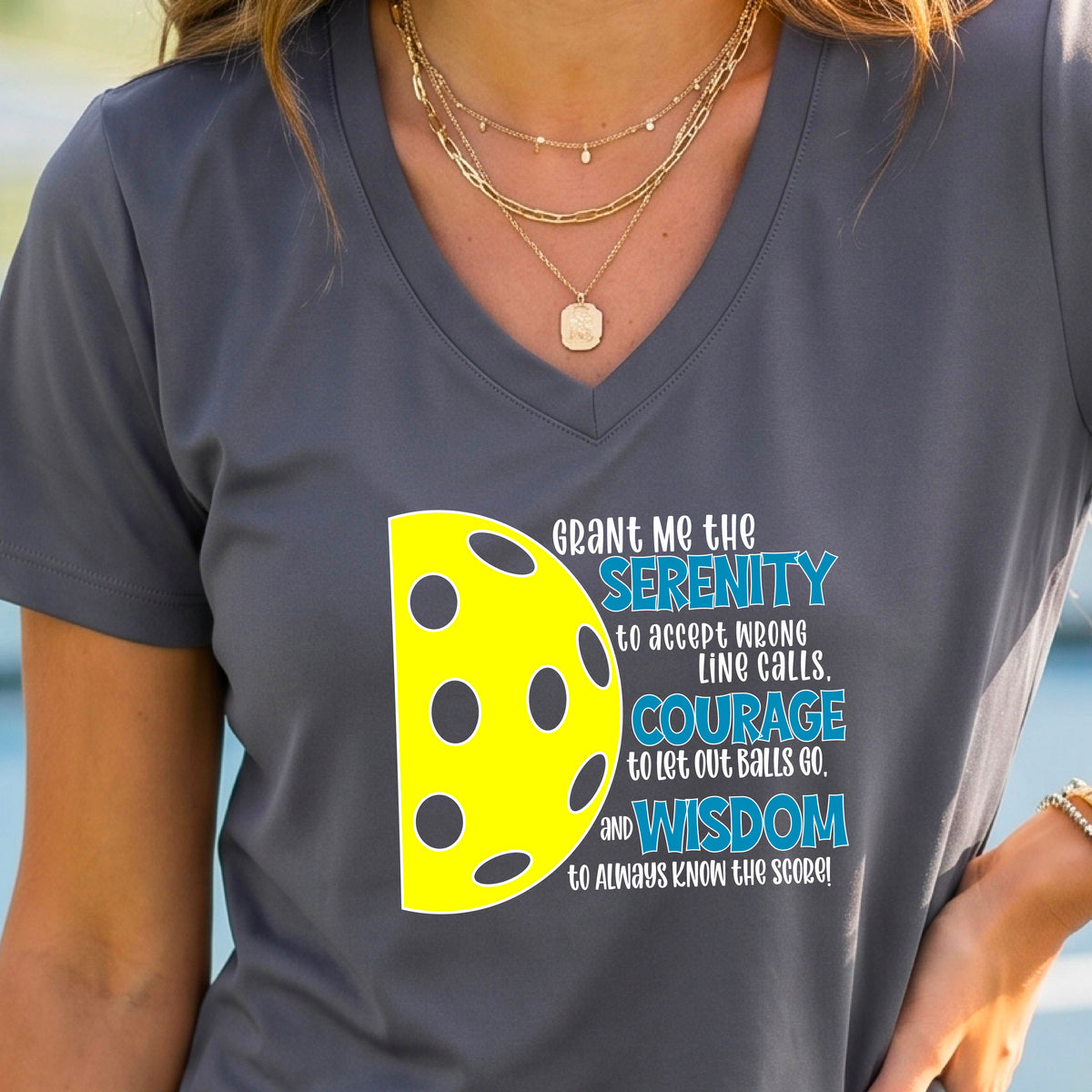 A woman on a pickleball court wearing a dark gray short-sleeve v-neck t-shirt with a graphic of the Serenity Prayer made for pickleball:  “Grant me the serenity to Accept Wrong Line Calls; Courage to Let Out Balls Go; and, Wisdom to Always Know the Score” next to a half-pickleball icon, standing on a pickleball court.