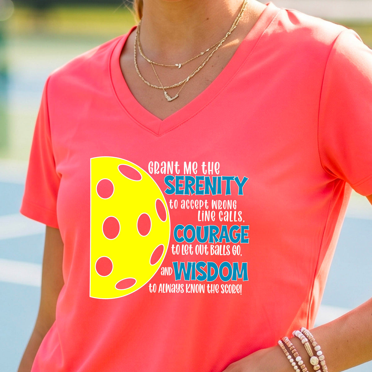 A woman on a pickleball court wearing a coral pink short-sleeve v-neck t-shirt with a graphic of the Serenity Prayer made for pickleball:  “Grant me the serenity to Accept Wrong Line Calls; Courage to Let Out Balls Go; and, Wisdom to Always Know the Score” next to a half-pickleball icon, standing on a pickleball court.