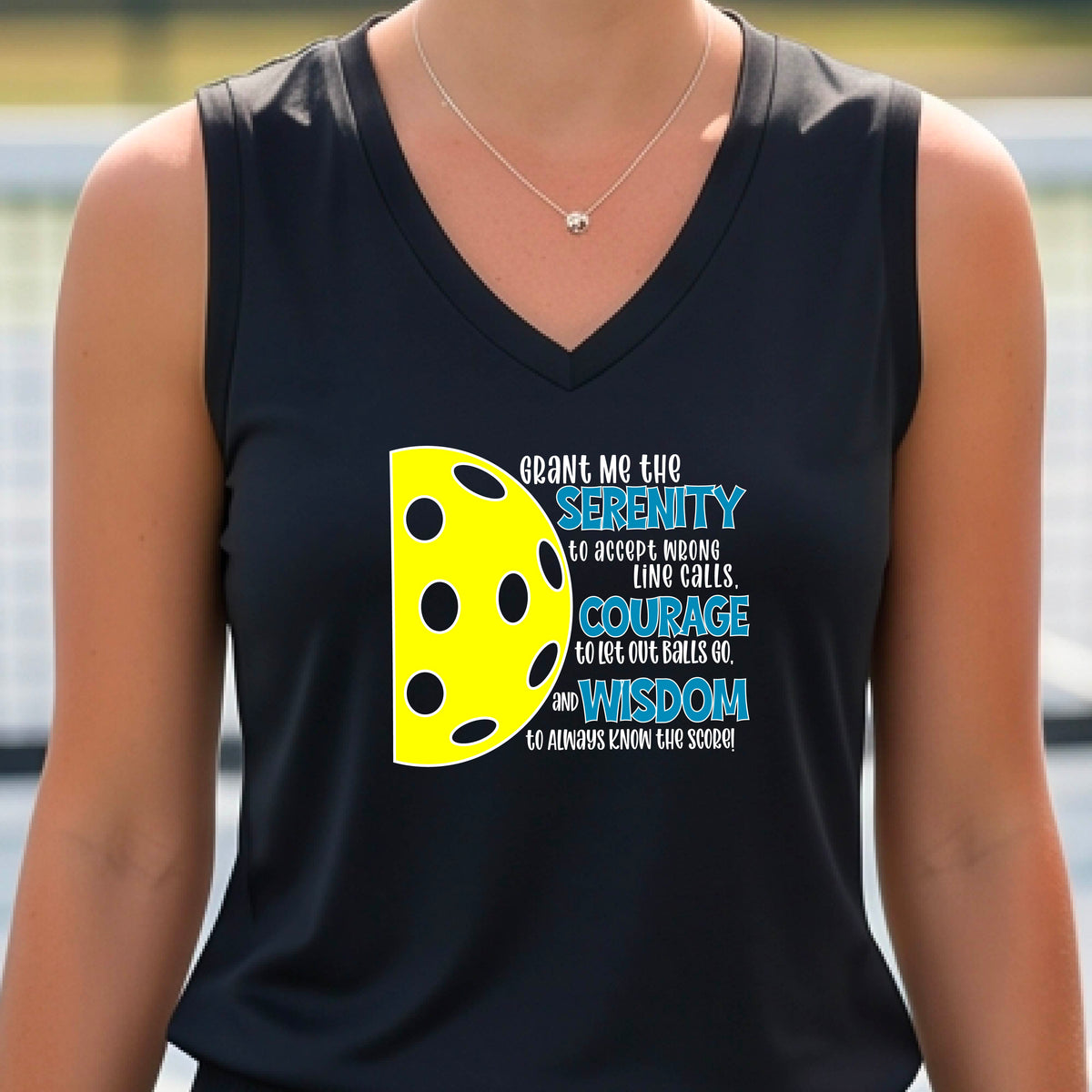 A woman on a pickleball court wearing a black sleeveless t-shirt with a graphic of the Serenity Prayer made for pickleball:  “Grant me the serenity to Accept Wrong Line Calls; Courage to Let Out Balls Go; and, Wisdom to Always Know the Score” next to a half-pickleball icon, standing on a pickleball court.