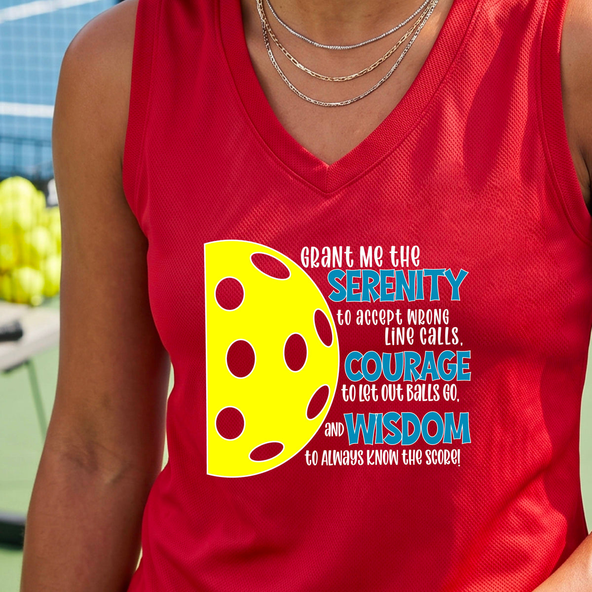 A woman on a pickleball court wearing a red sleeveless t-shirt with a graphic of the Serenity Prayer made for pickleball:  “Grant me the serenity to Accept Wrong Line Calls; Courage to Let Out Balls Go; and, Wisdom to Always Know the Score” next to a half-pickleball icon, standing on a pickleball court.