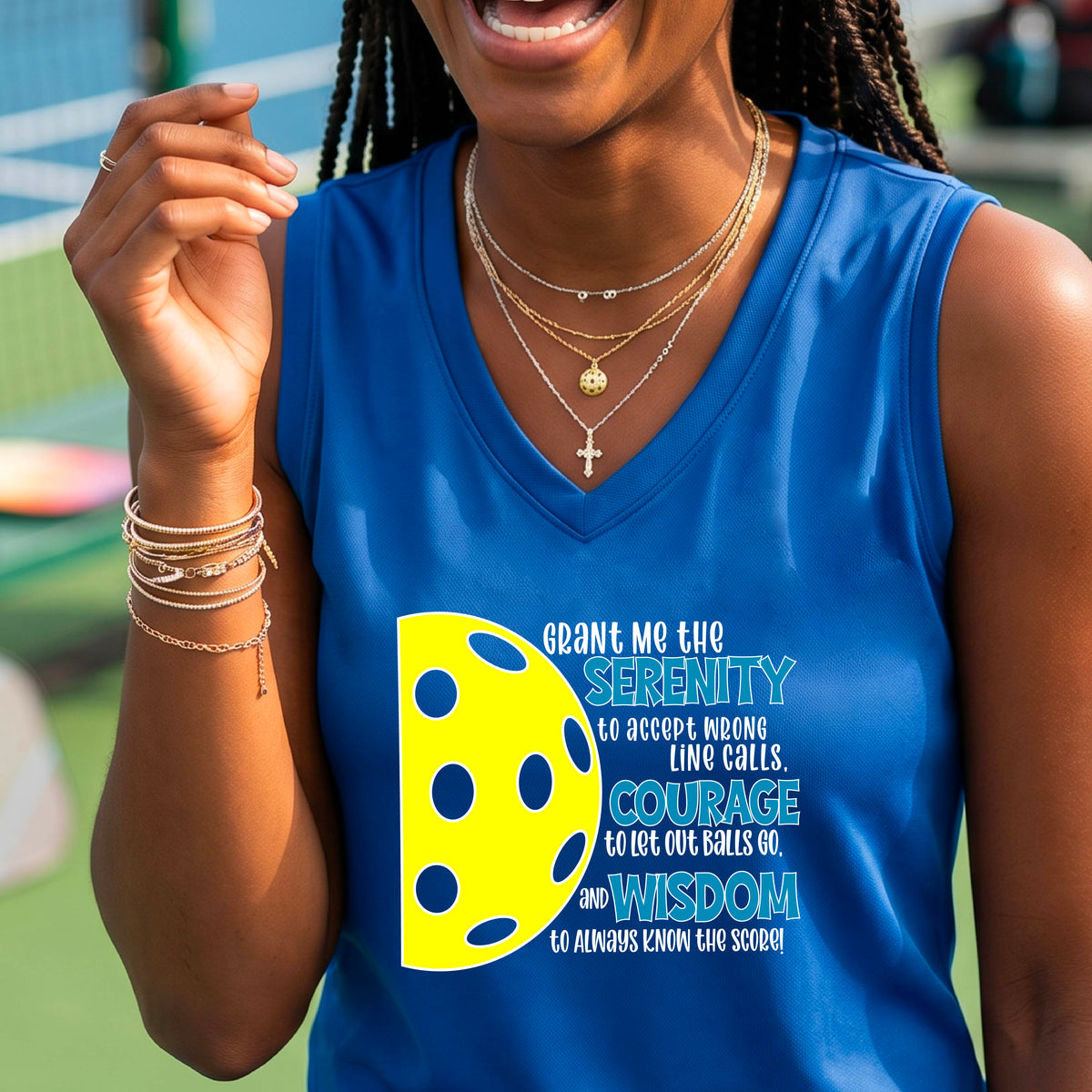 A woman on a pickleball court wearing a royal blue sleeveless t-shirt with a graphic of the Serenity Prayer made for pickleball:  “Grant me the serenity to Accept Wrong Line Calls; Courage to Let Out Balls Go; and, Wisdom to Always Know the Score” next to a half-pickleball icon, standing on a pickleball court.