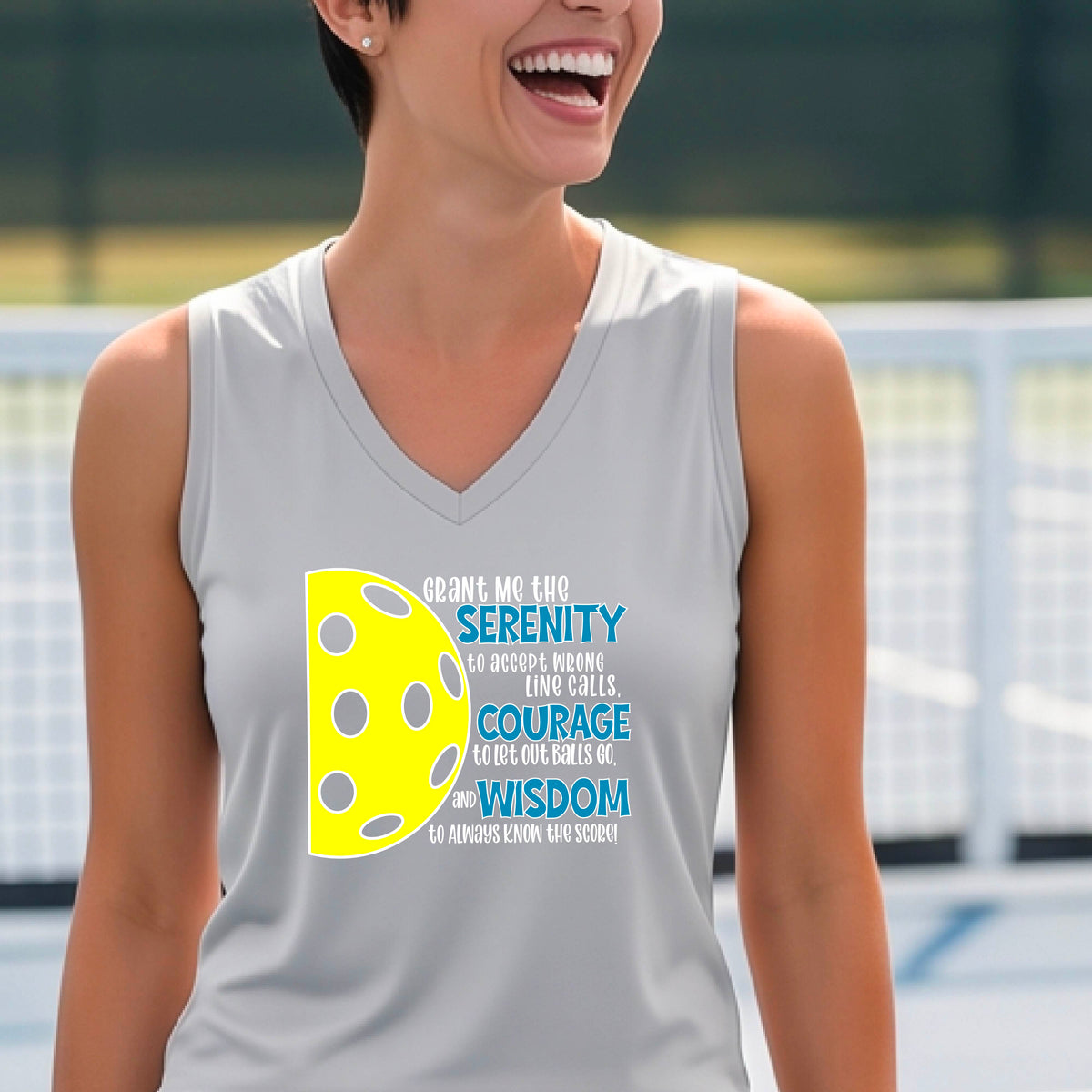 A woman on a pickleball court wearing a gray sleeveless t-shirt with a graphic of the Serenity Prayer made for pickleball:  “Grant me the serenity to Accept Wrong Line Calls; Courage to Let Out Balls Go; and, Wisdom to Always Know the Score” next to a half-pickleball icon, standing on a pickleball court.