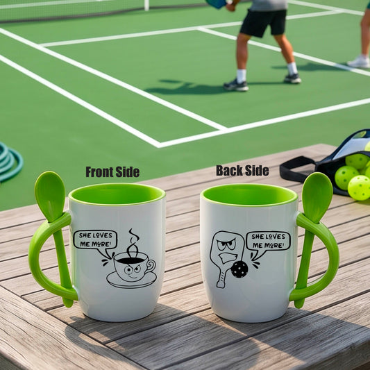 A white and green 12 ounce coffee mug with a built in spoon in the handle with and a fun pickleball paddle and ball and coffee cup graphic and the text “SHE LOVES ME MORE”