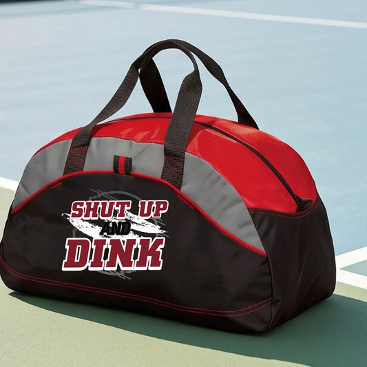 A close-up of a red and black with gray panels, custom duffel bag with a white, gray and black graphic of the text: ‘Shut Up and Dink, standing on a pickleball court.