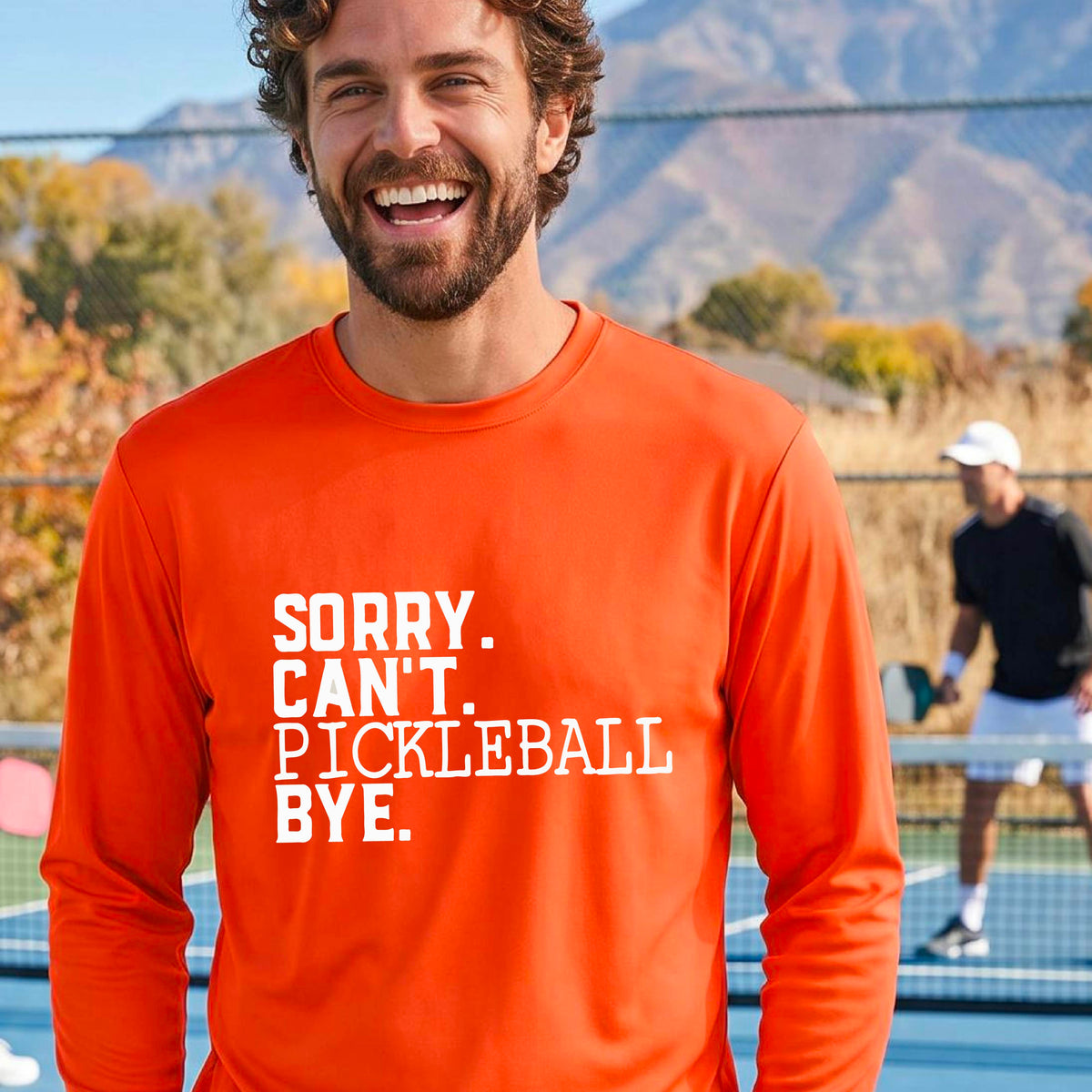 A man on a pickleball court wearing a bright orange long-sleeve t-shirt with a white graphic of the words  “SORRY. CAN’T. PICKLEBALL BYE” each on a separate line, on the front of the shirt. 