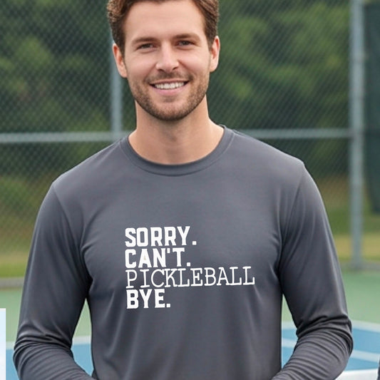 A man on a pickleball court wearing a gray long-sleeve t-shirt with a white graphic of the words  “SORRY. CAN’T. PICKLEBALL BYE” each on a separate line, on the front of the shirt. 