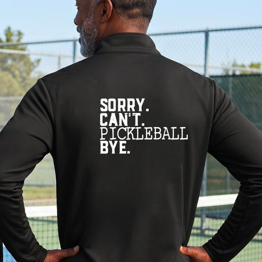 A man viewed from behind on a pickleball court wearing a black quarter-zip shirt with a white graphic of the words  “SORRY. CAN’T. PICKLEBALL BYE” each on a separate line, on the front of the shirt.