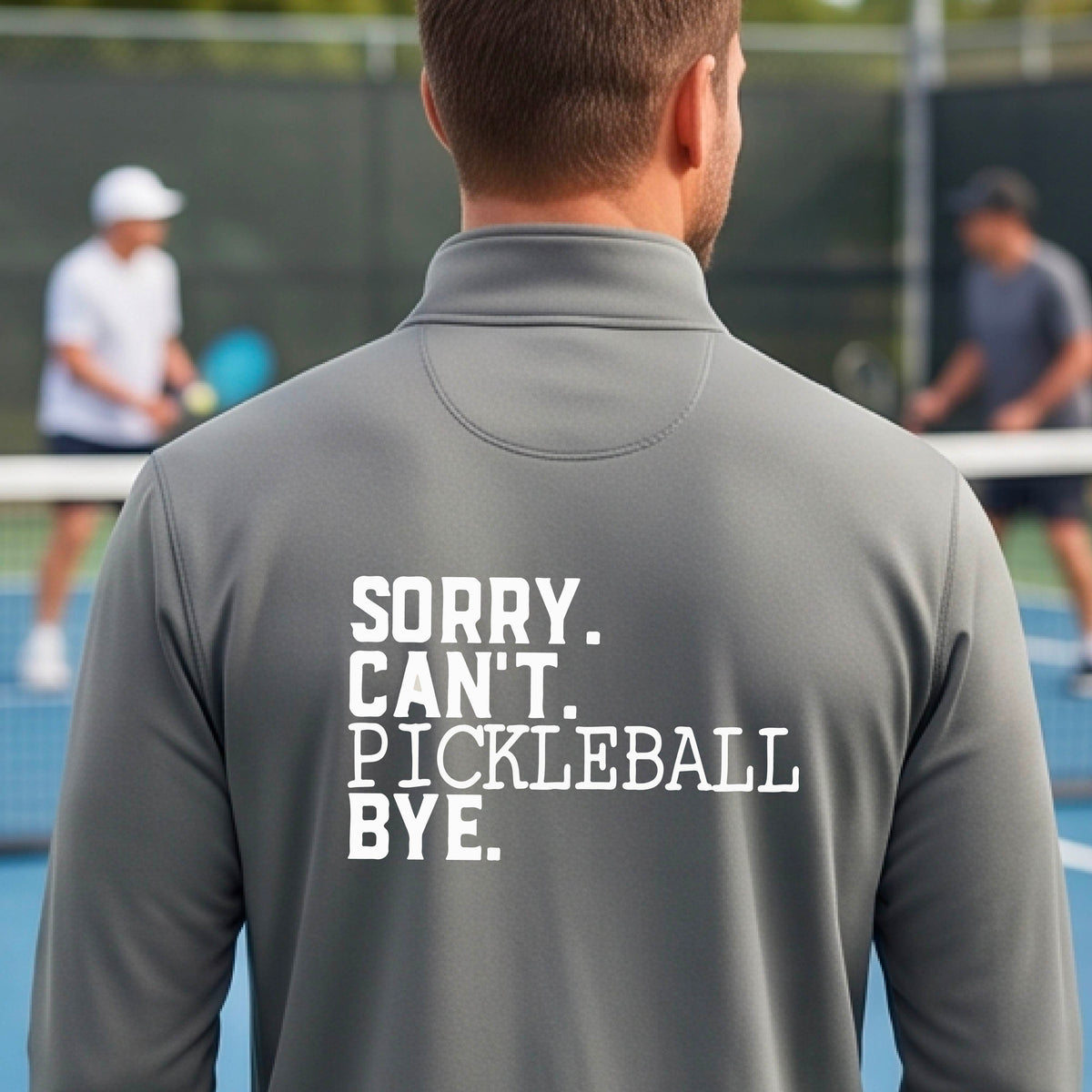 A man viewed from behind on a pickleball court wearing a gray quarter-zip shirt with a white graphic of the words  “SORRY. CAN’T. PICKLEBALL BYE” each on a separate line, on the front of the shirt.