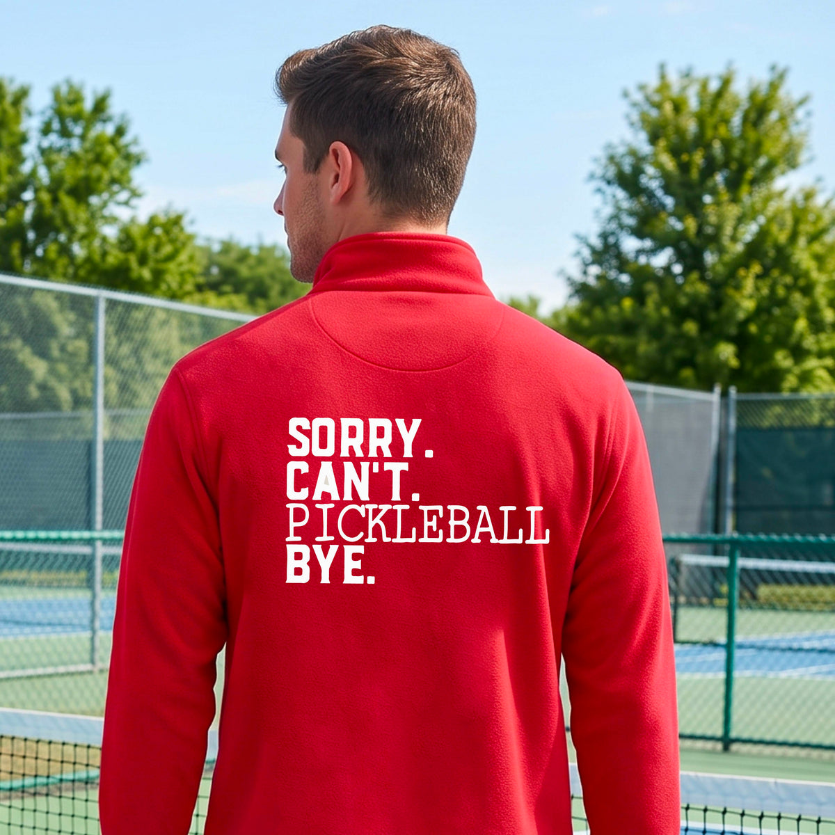 A man viewed from behind on a pickleball court wearing a red quarter-zip shirt with a white graphic of the words  “SORRY. CAN’T. PICKLEBALL BYE” each on a separate line, on the front of the shirt.