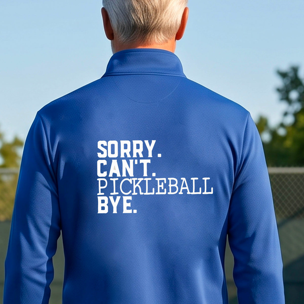 A man viewed from behind on a pickleball court wearing a royal blue quarter-zip shirt with a white graphic of the words  “SORRY. CAN’T. PICKLEBALL BYE” each on a separate line, on the front of the shirt.