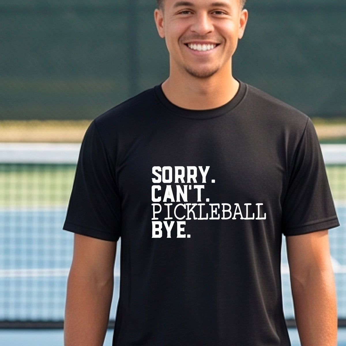 A man on a pickleball court wearing a black short-sleeve t-shirt with a white graphic of the words  “SORRY. CAN’T. PICKLEBALL BYE” each on a separate line, on the front of the shirt. 