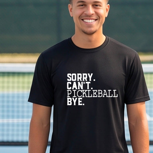 A man on a pickleball court wearing a black short-sleeve t-shirt with a white graphic of the words  “SORRY. CAN’T. PICKLEBALL BYE” each on a separate line, on the front of the shirt. 
