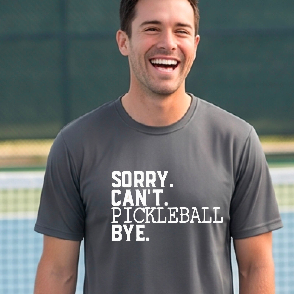 A man on a pickleball court wearing a gray short-sleeve t-shirt with a white graphic of the words  “SORRY. CAN’T. PICKLEBALL BYE” each on a separate line, on the front of the shirt. 