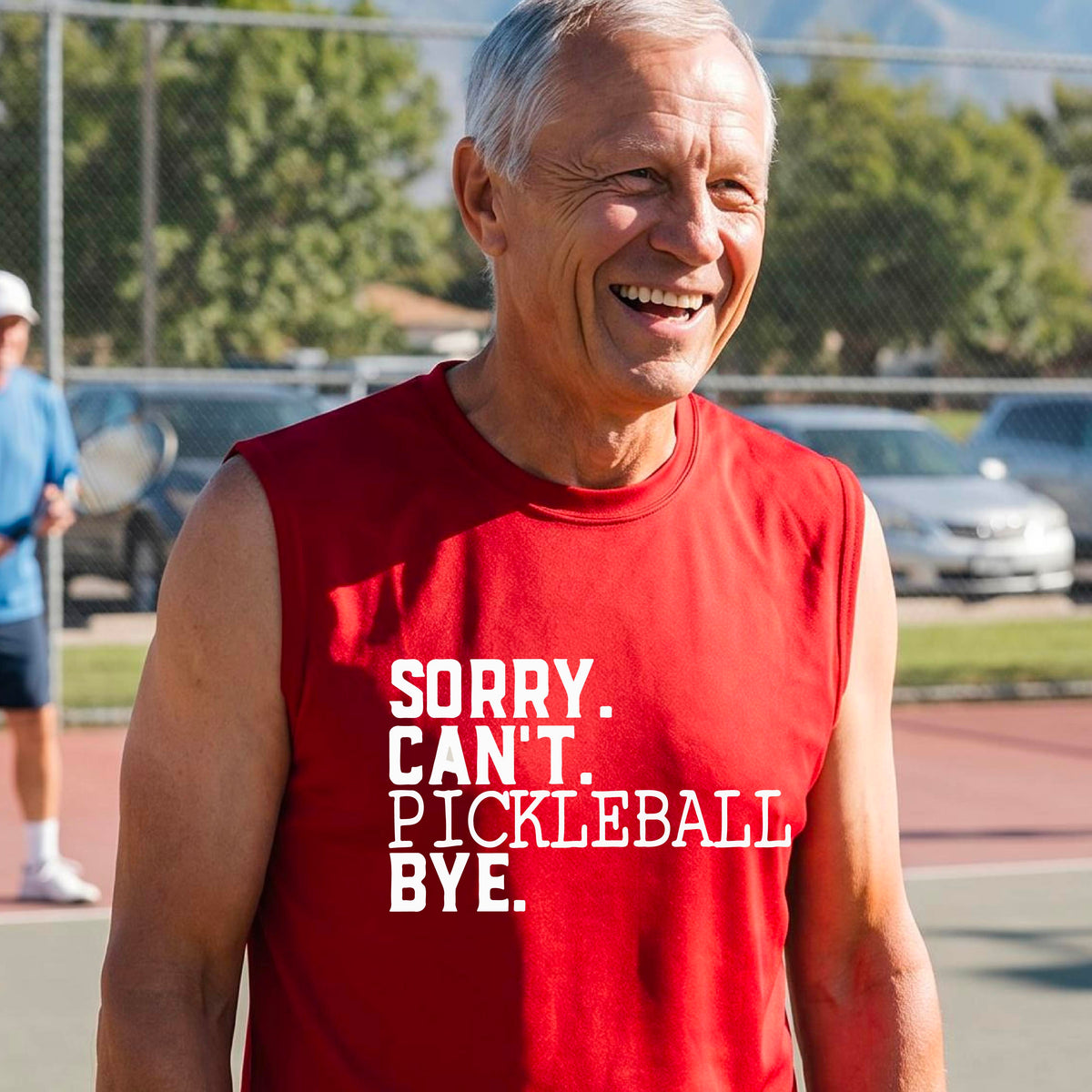 A man on a pickleball court wearing a red sleeveless t-shirt with a white graphic of the words  “SORRY. CAN’T. PICKLEBALL BYE” each on a separate line, on the front of the shirt. 