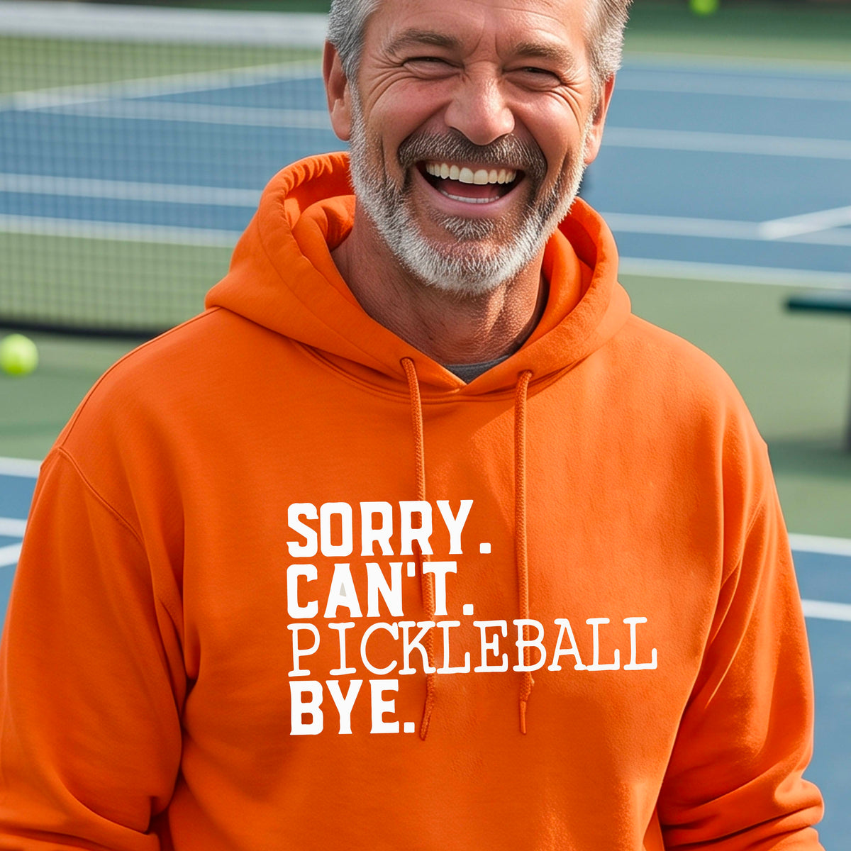 A man on a pickleball court wearing an orange unisex hoodie pullover with a white graphic of the words  “SORRY. CAN’T. PICKLEBALL BYE” each on a separate line, on the front of the shirt. 