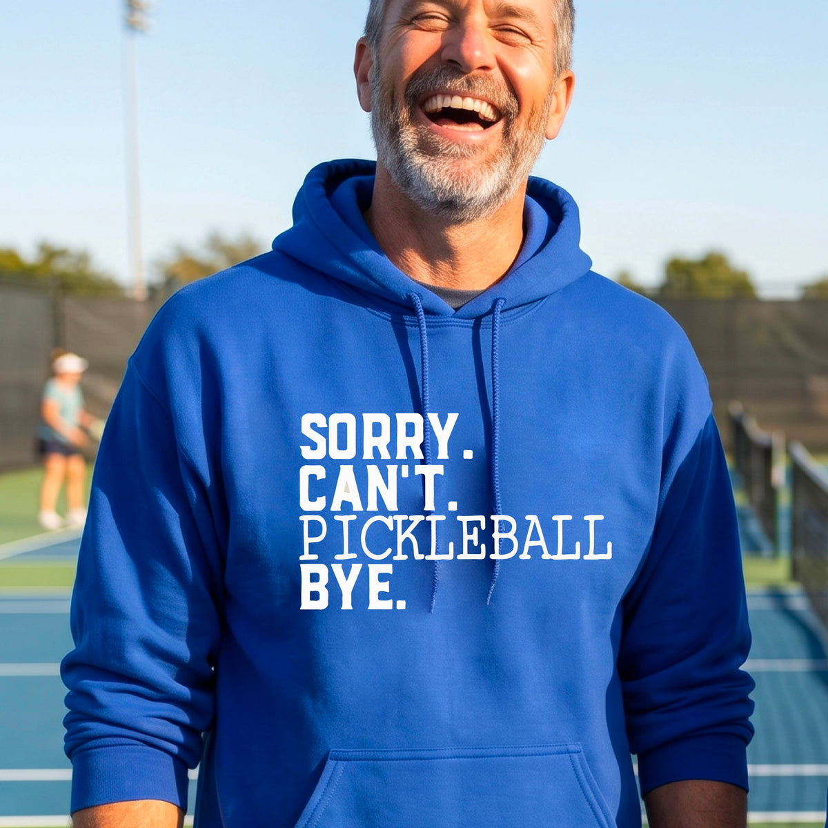 A man on a pickleball court wearing a royal blue unisex hoodie pullover with a white graphic of the words  “SORRY. CAN’T. PICKLEBALL BYE” each on a separate line, on the front of the shirt. 