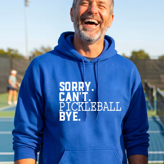 A man on a pickleball court wearing a royal blue unisex hoodie pullover with a white graphic of the words  “SORRY. CAN’T. PICKLEBALL BYE” each on a separate line, on the front of the shirt. 
