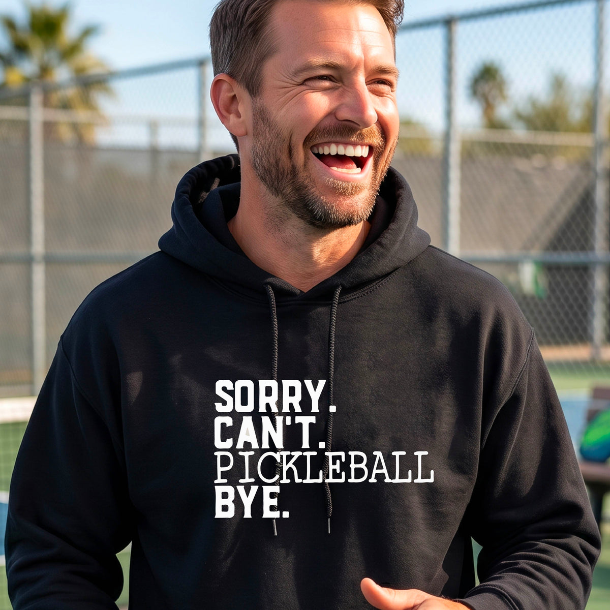A man on a pickleball court wearing a black unisex hoodie pullover with a white graphic of the words  “SORRY. CAN’T. PICKLEBALL BYE” each on a separate line, on the front of the shirt. 