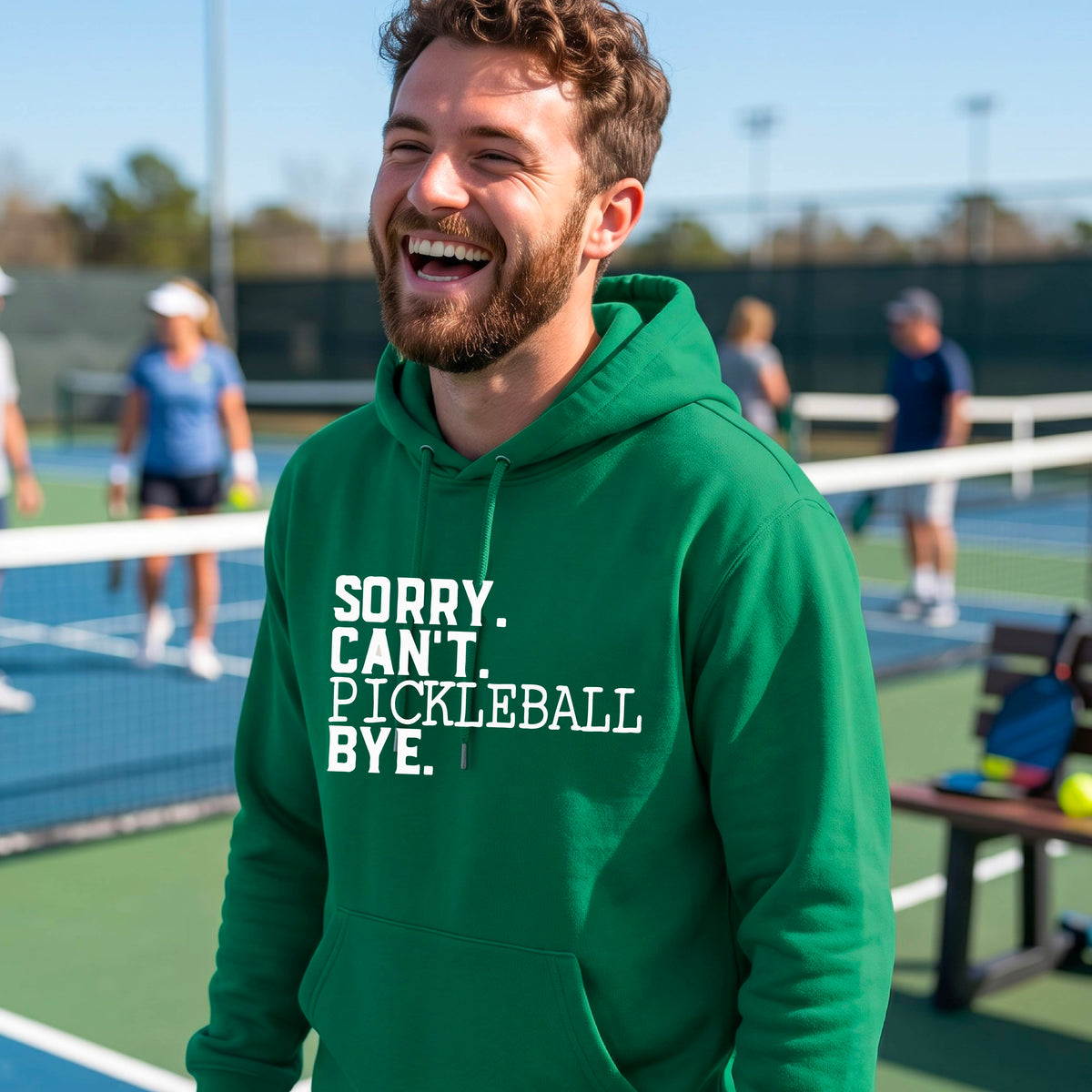 A man on a pickleball court wearing a green unisex hoodie pullover with a white graphic of the words  “SORRY. CAN’T. PICKLEBALL BYE” each on a separate line, on the front of the shirt. 