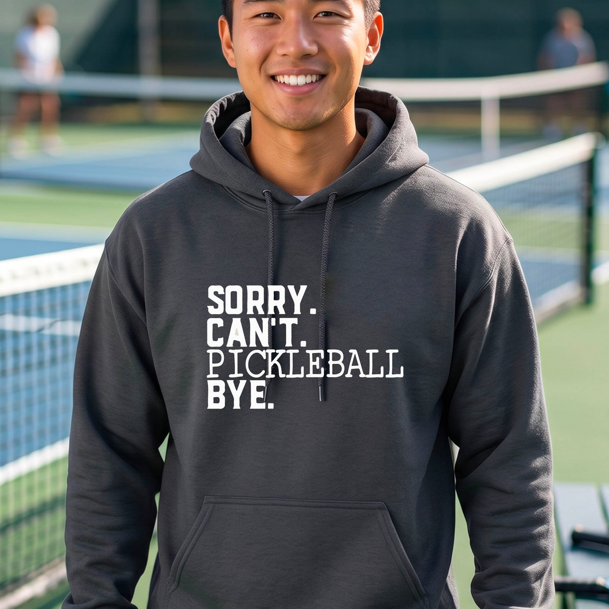 A man on a pickleball court wearing a dark gray unisex hoodie pullover with a white graphic of the words  “SORRY. CAN’T. PICKLEBALL BYE” each on a separate line, on the front of the shirt. 