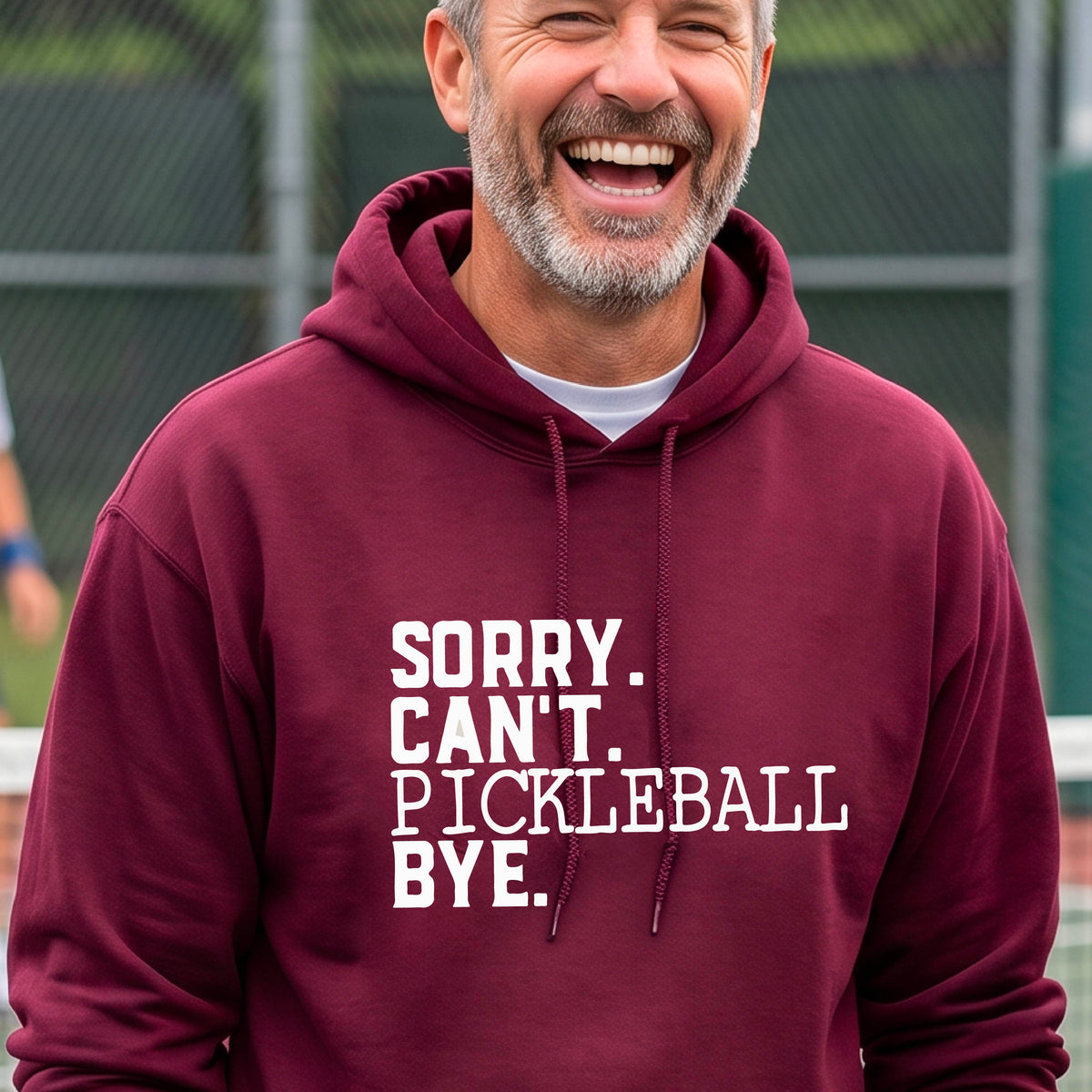 A man on a pickleball court wearing a maroon unisex hoodie pullover with a white graphic of the words  “SORRY. CAN’T. PICKLEBALL BYE” each on a separate line, on the front of the shirt. 