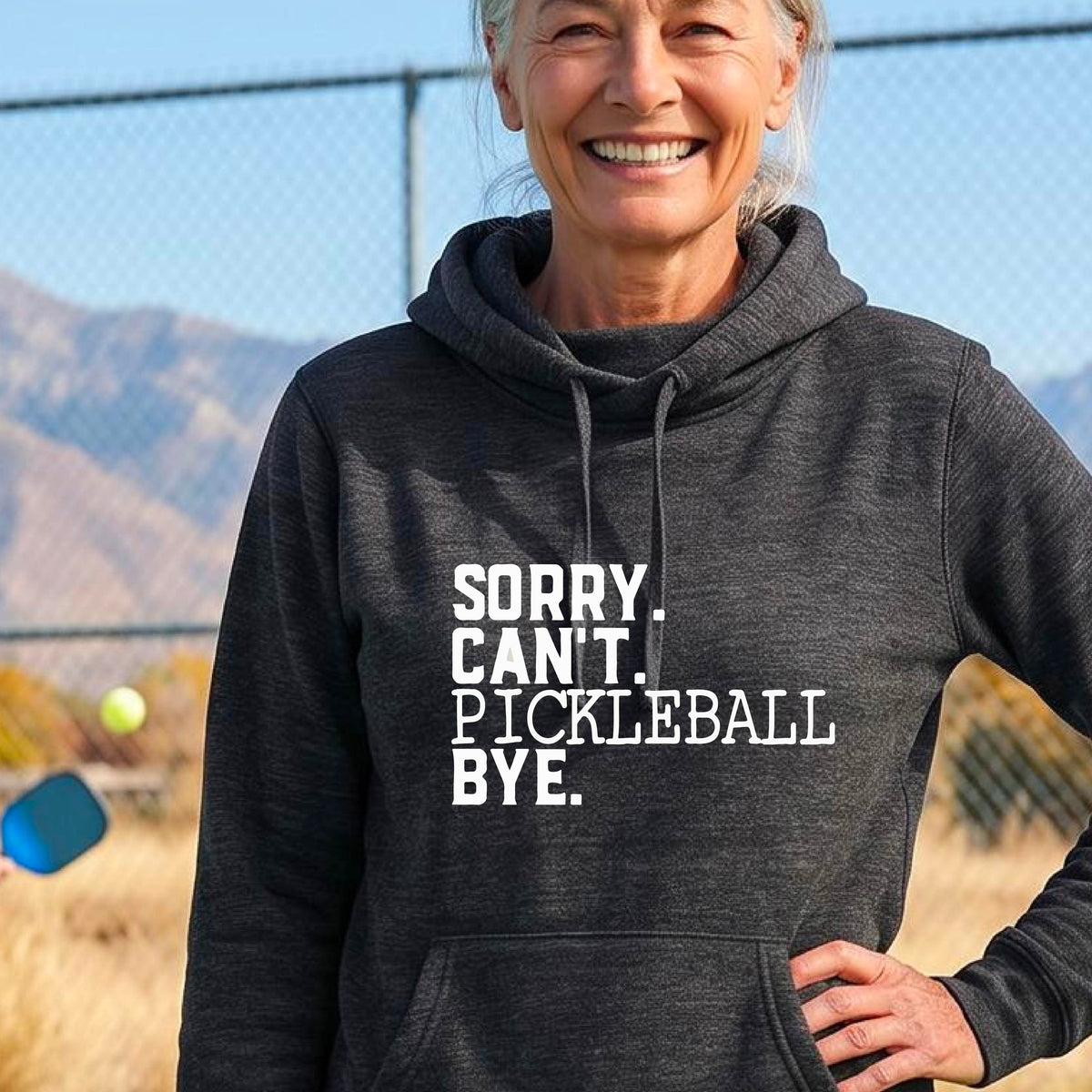 A woman on a pickleball court wearing a black cowl-neck hoodie pullover with a white graphic of the words  “SORRY. CAN’T. PICKLEBALL BYE” each on a separate line, on the front. 