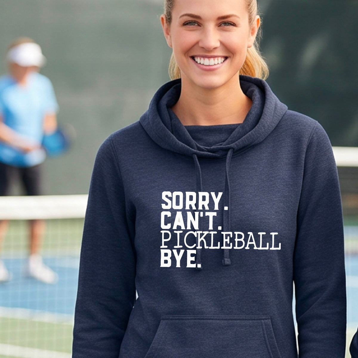 A woman on a pickleball court wearing a navy blue cowl-neck hoodie pullover with a white graphic of the words  “SORRY. CAN’T. PICKLEBALL BYE” each on a separate line, on the front. 
