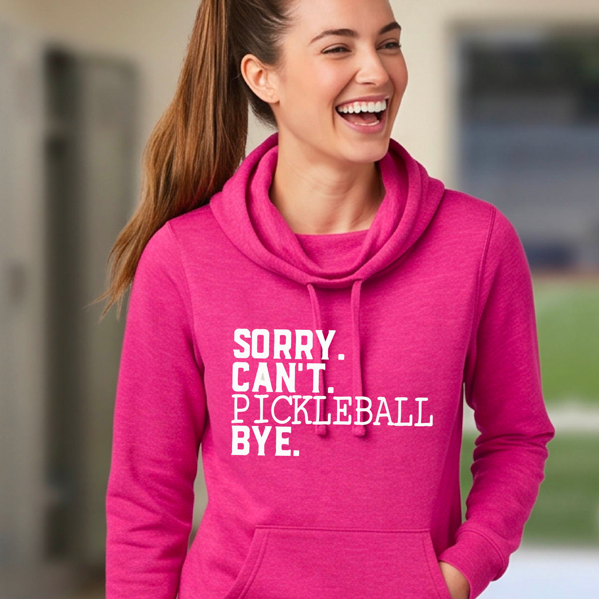A woman on a pickleball court wearing a pink cowl-neck hoodie pullover with a white graphic of the words  “SORRY. CAN’T. PICKLEBALL BYE” each on a separate line, on the front. 