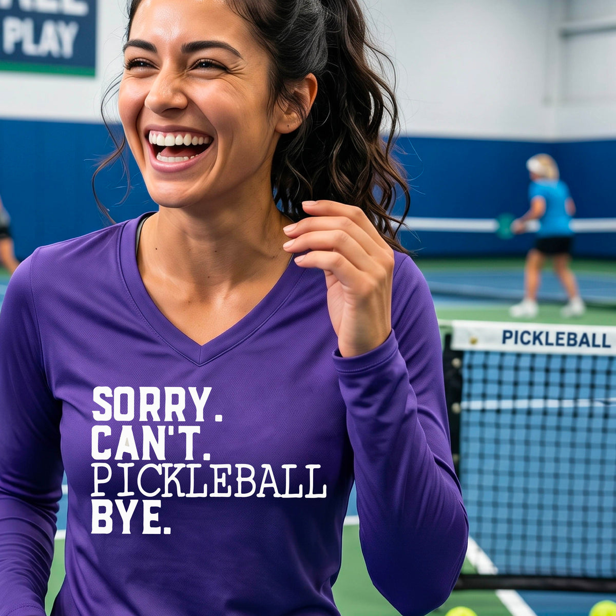 A woman on a pickleball court wearing a purple long-sleeve t-shirt with a white graphic of the words  “SORRY. CAN’T. PICKLEBALL BYE” each on a separate line, on the front of the shirt. 