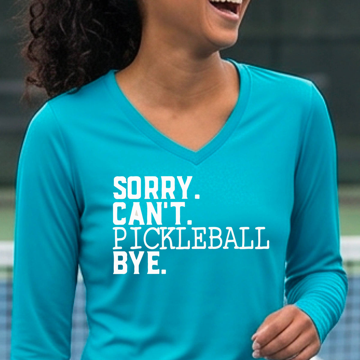 A woman on a pickleball court wearing a bright blue long-sleeve t-shirt with a white graphic of the words  “SORRY. CAN’T. PICKLEBALL BYE” each on a separate line, on the front of the shirt. 