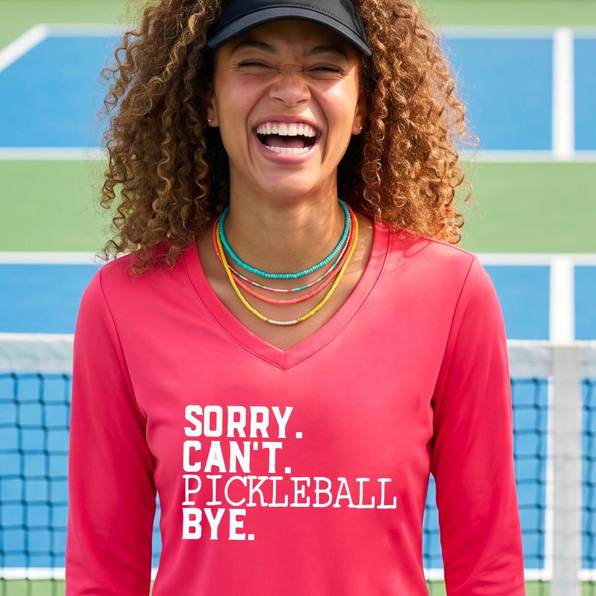 A woman on a pickleball court wearing a coral red long-sleeve t-shirt with a white graphic of the words  “SORRY. CAN’T. PICKLEBALL BYE” each on a separate line, on the front of the shirt. 