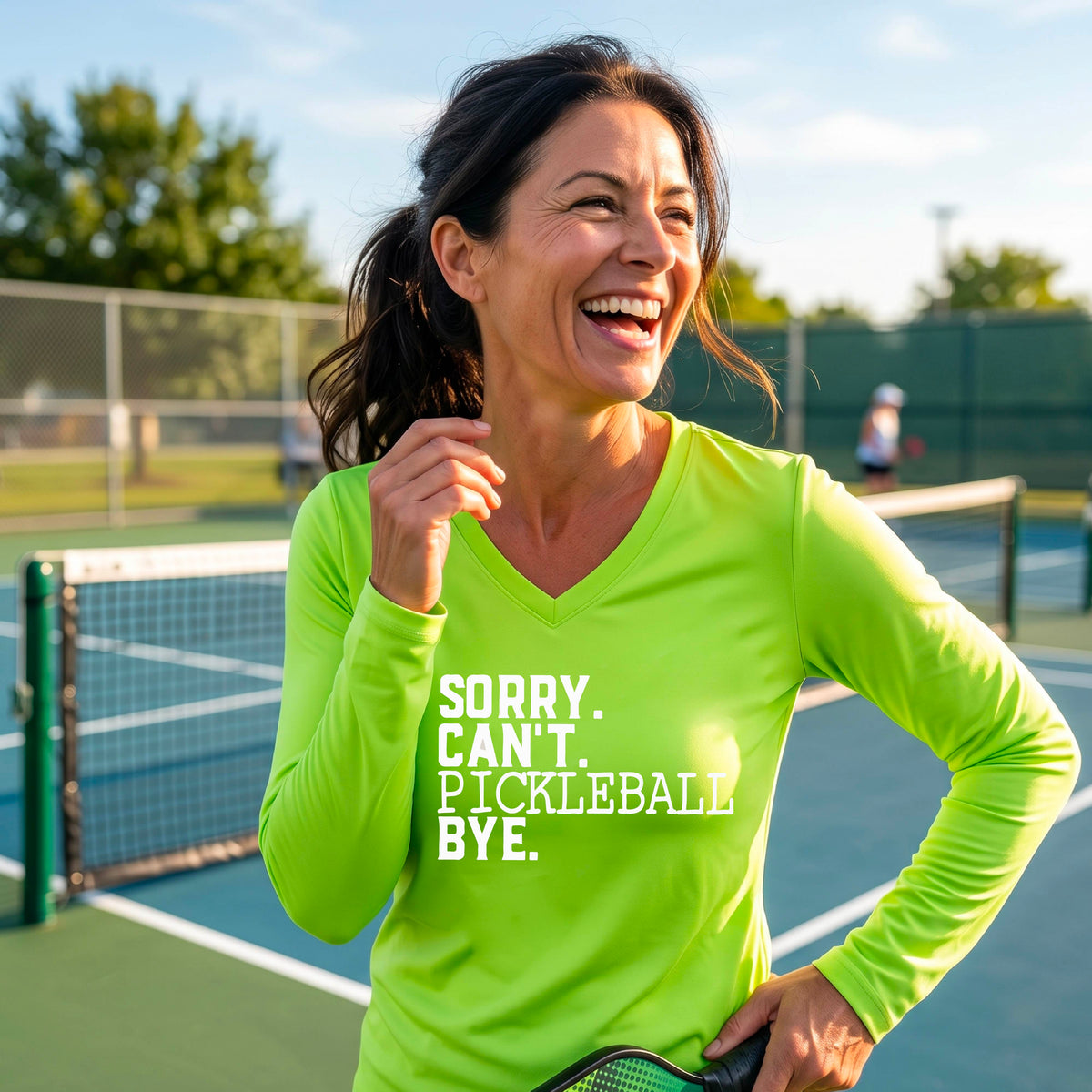A woman on a pickleball court wearing a lime green long-sleeve t-shirt with a white graphic of the words  “SORRY. CAN’T. PICKLEBALL BYE” each on a separate line, on the front of the shirt. 