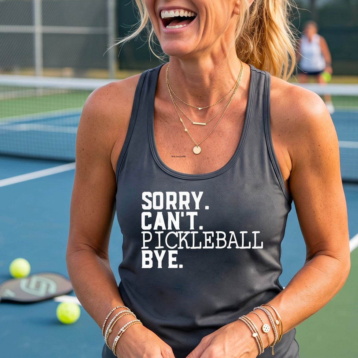 A woman on a pickleball court wearing a graphite gray racerback tank top with a white graphic of the words  “SORRY. CAN’T. PICKLEBALL BYE” each on a separate line, on the fron of the shirt. 