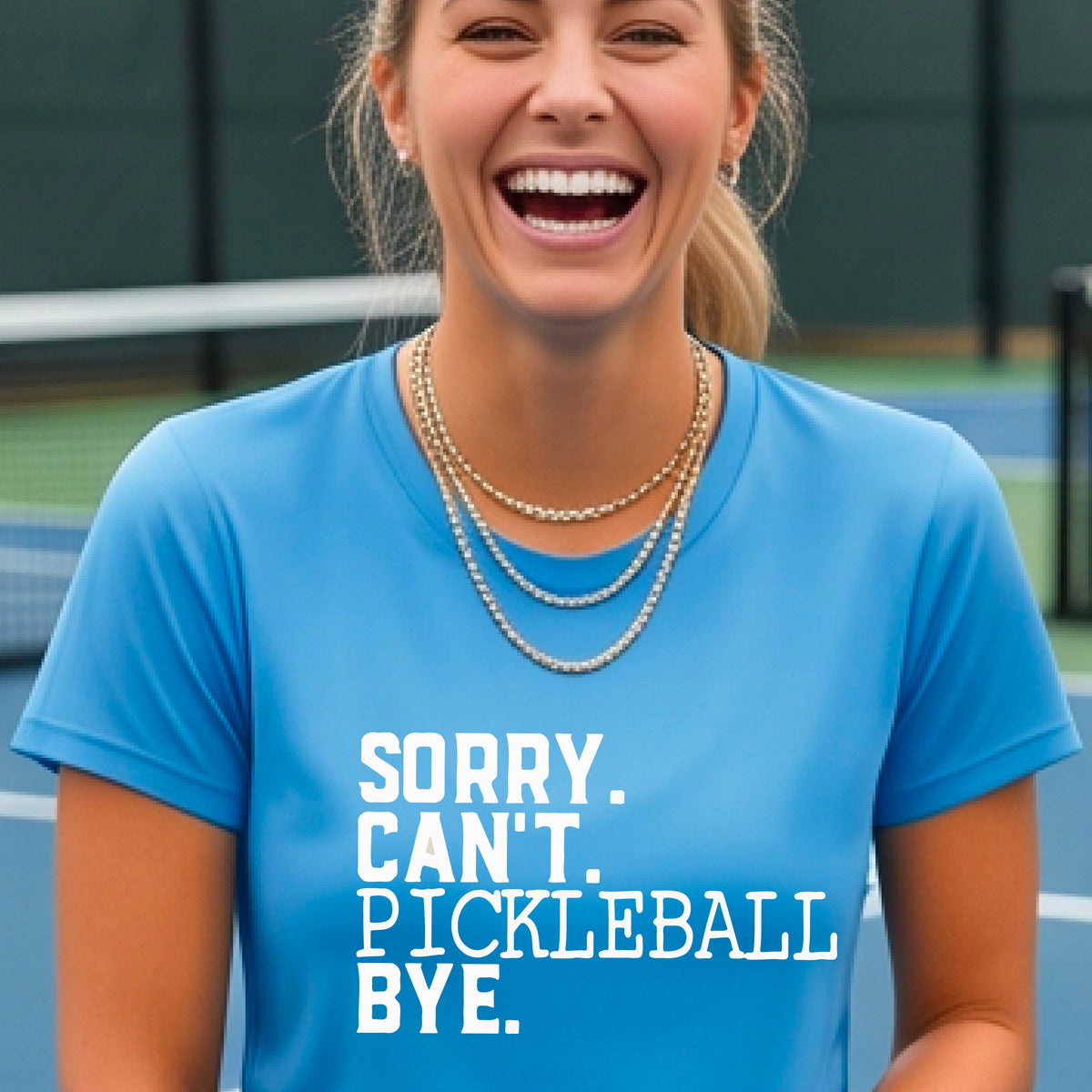 A woman on a pickleball court wearing a blue short-sleeve t-shirt with a white graphic of the words  “SORRY. CAN’T. PICKLEBALL BYE” each on a separate line, on the front of the shirt. 

