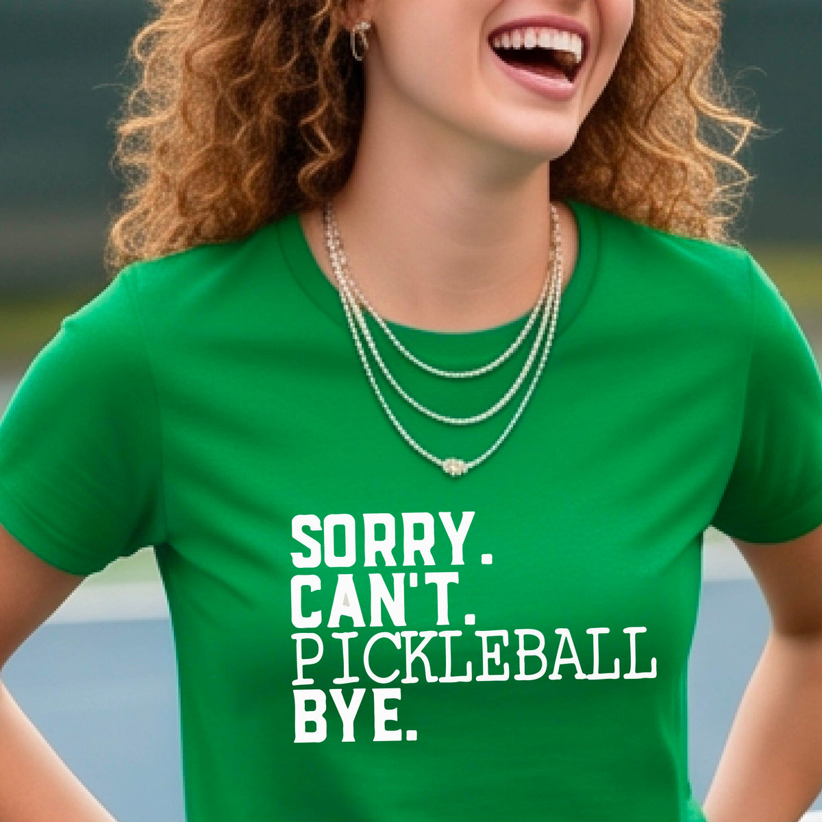 A woman on a pickleball court wearing a green short-sleeve t-shirt with a white graphic of the words  “SORRY. CAN’T. PICKLEBALL BYE” each on a separate line, on the front of the shirt. 

