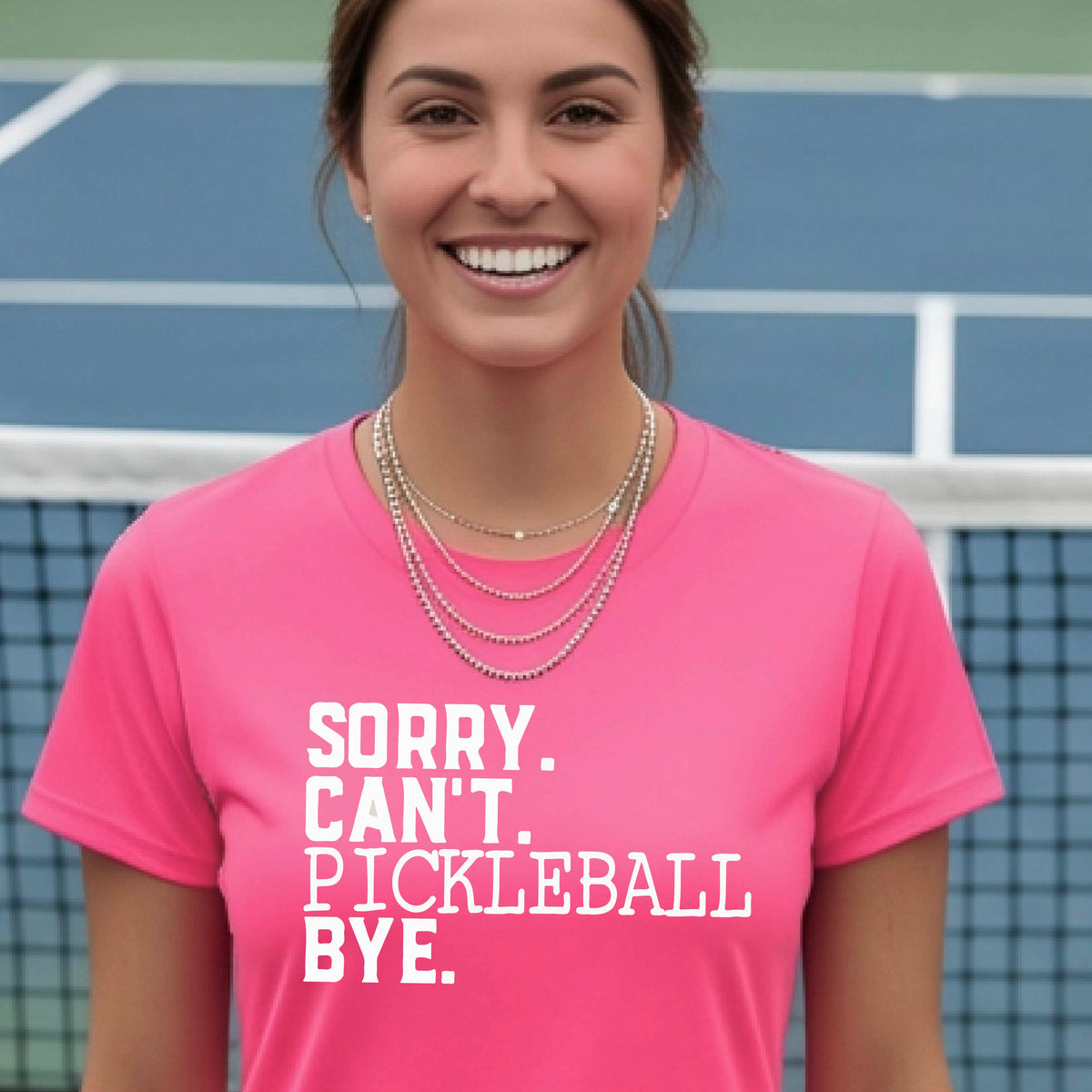 A woman on a pickleball court wearing a pink short-sleeve t-shirt with a white graphic of the words  “SORRY. CAN’T. PICKLEBALL BYE” each on a separate line, on the front of the shirt. 

