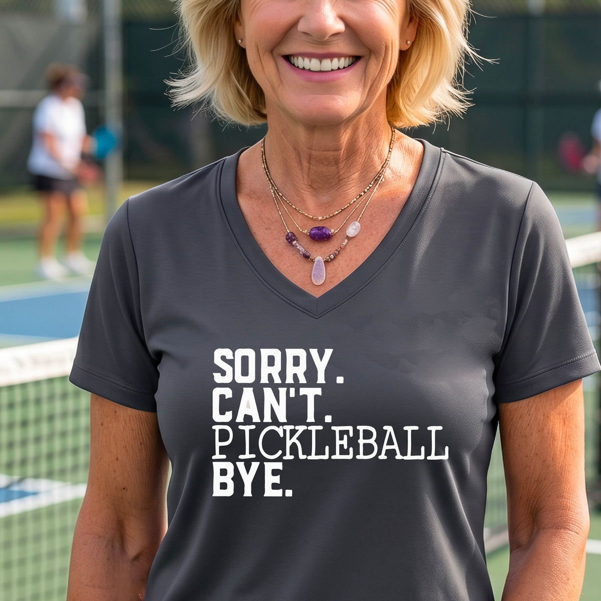 A woman on a pickleball court wearing a gray short-sleeve t-shirt with a white graphic of the words  “SORRY. CAN’T. PICKLEBALL BYE” each on a separate line, on the front of the shirt. 