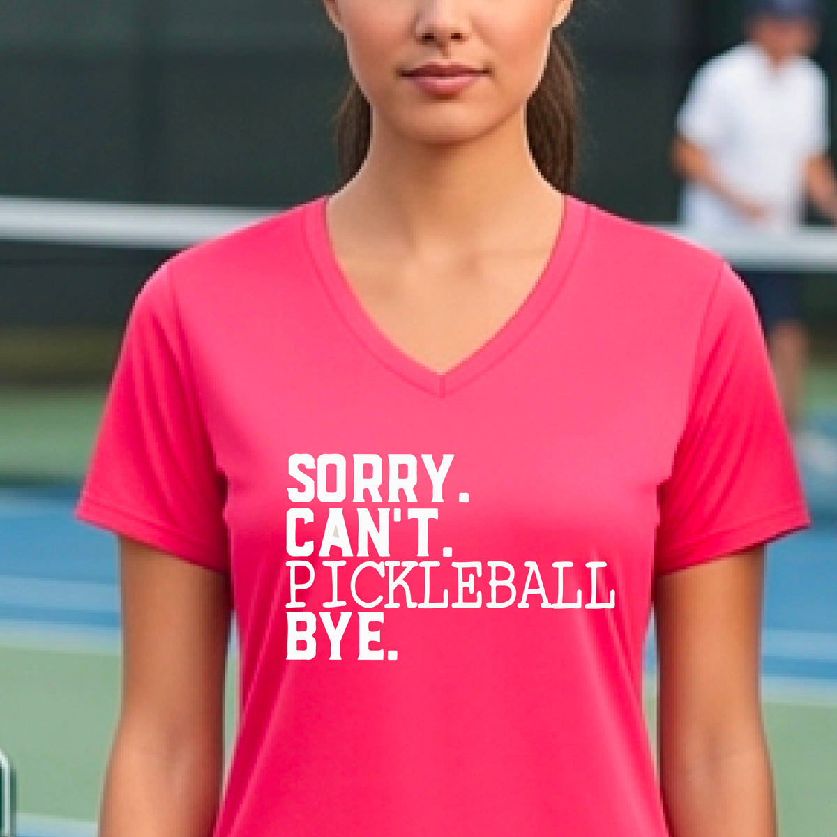 A woman on a pickleball court wearing a coral pink short-sleeve t-shirt with a white graphic of the words  “SORRY. CAN’T. PICKLEBALL BYE” each on a separate line, on the front of the shirt. 