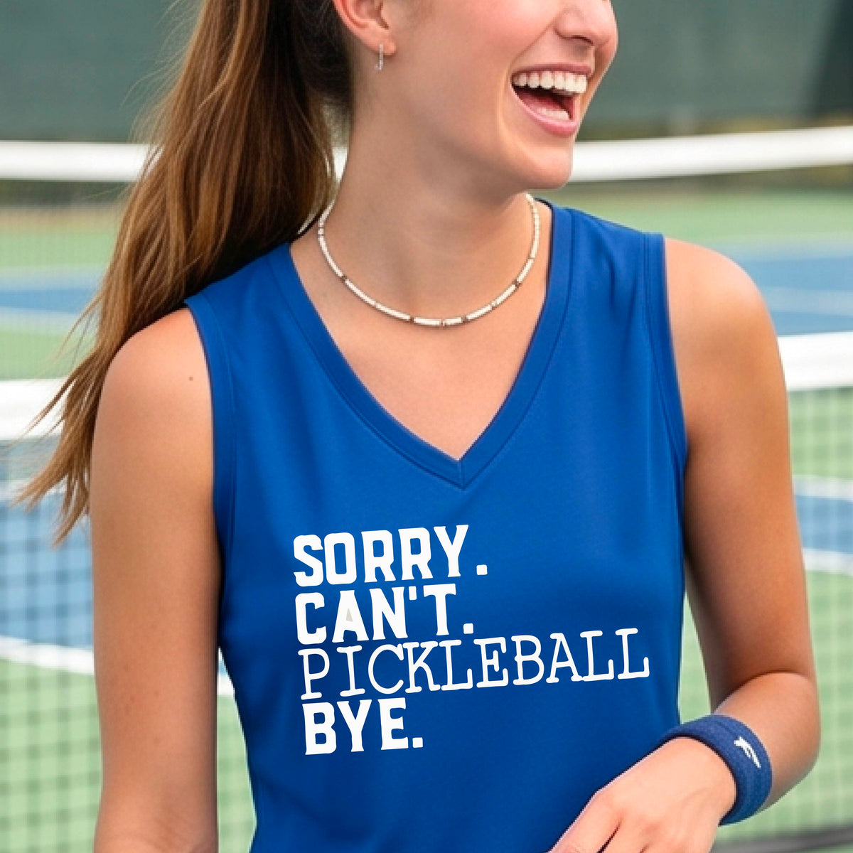 A woman on a pickleball court wearing a royal blue sleeveless t-shirt with a white graphic of the words  “SORRY. CAN’T. PICKLEBALL BYE” each on a separate line, on the front of the shirt. 