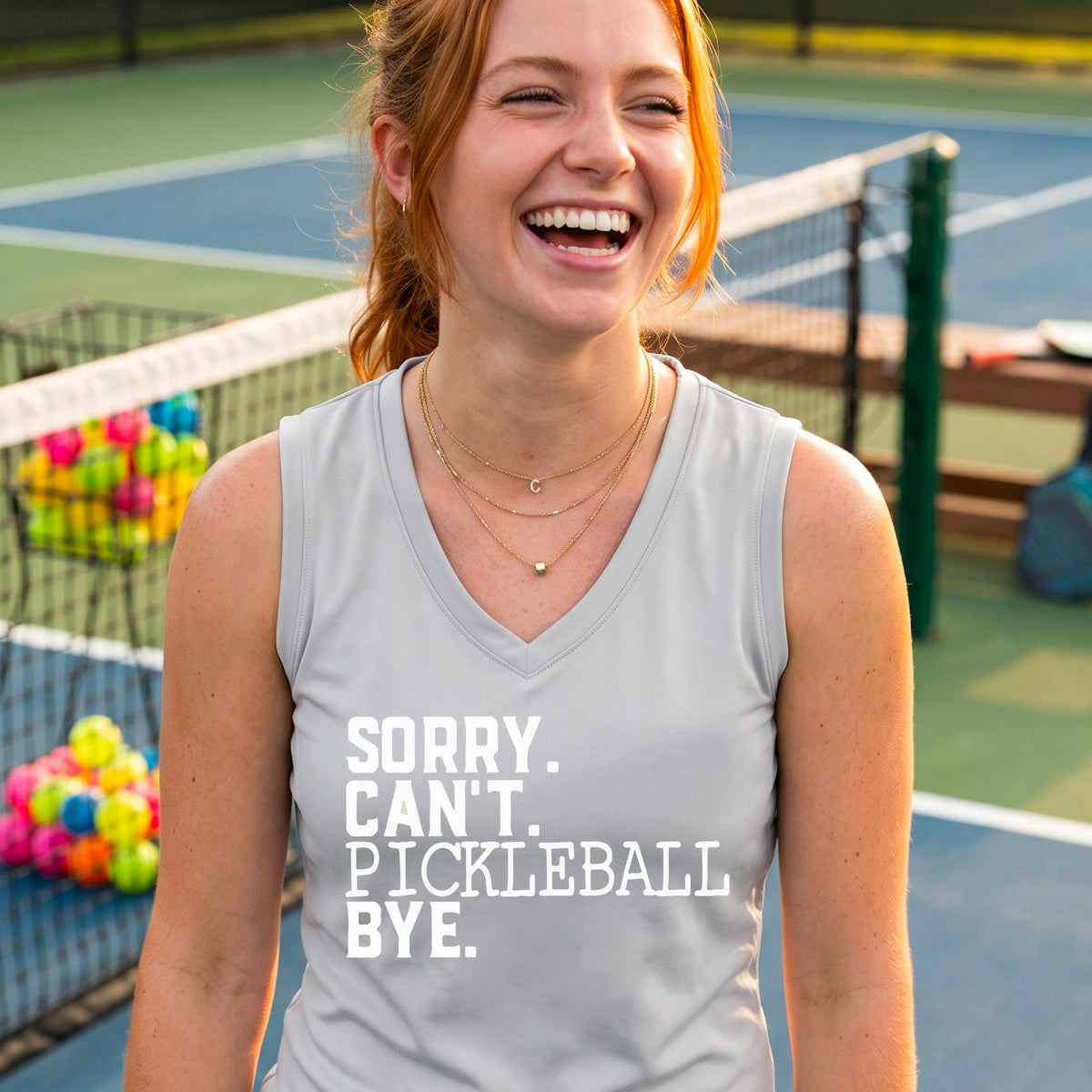 A woman on a pickleball court wearing a silver gray sleeveless t-shirt with a white graphic of the words  “SORRY. CAN’T. PICKLEBALL BYE” each on a separate line, on the front of the shirt. 