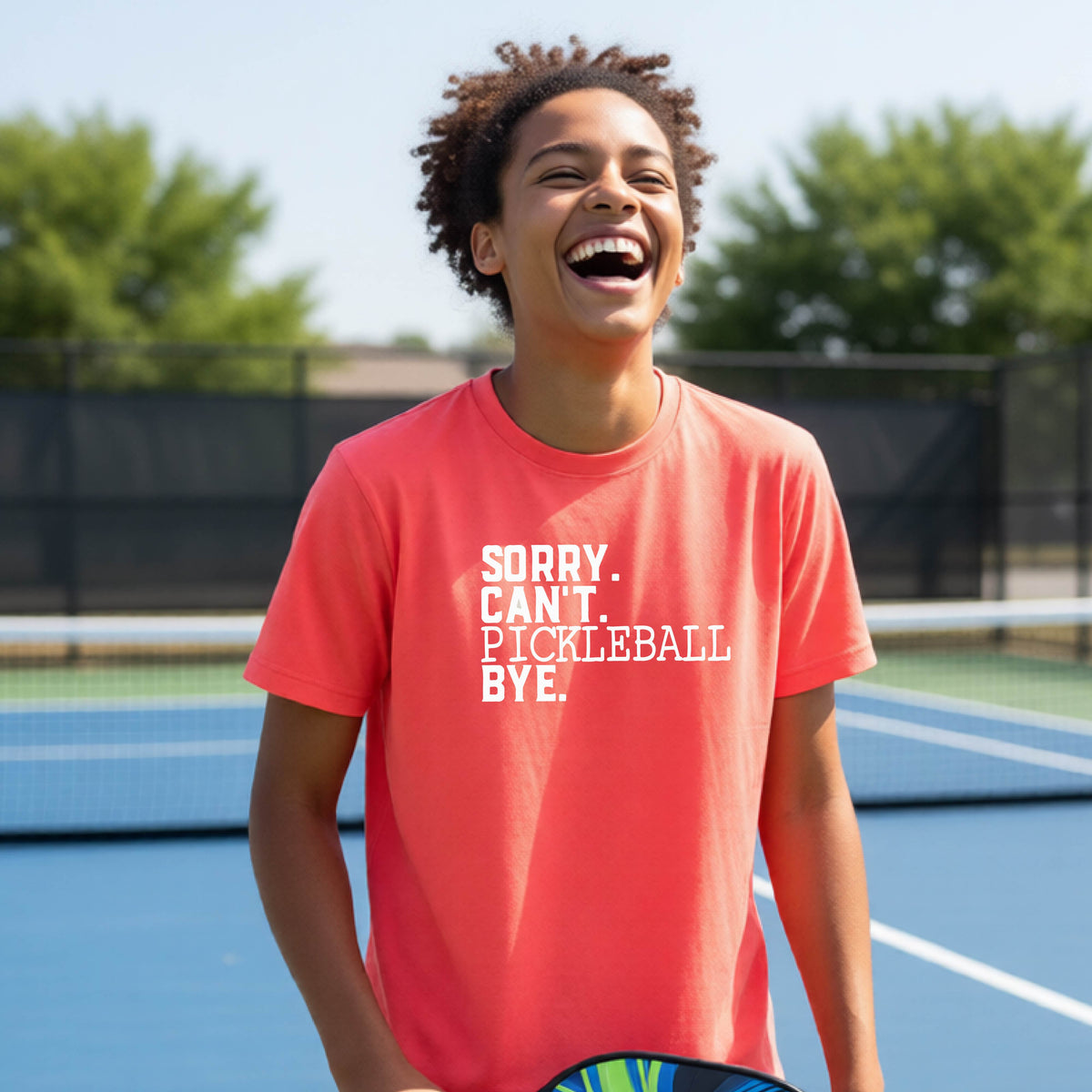 A young person on a pickleball court wearing a coral pink short-sleeve t-shirt with a white graphic of the words  “SORRY. CAN’T. PICKLEBALL BYE” each on a separate line, on the front of the shirt. 