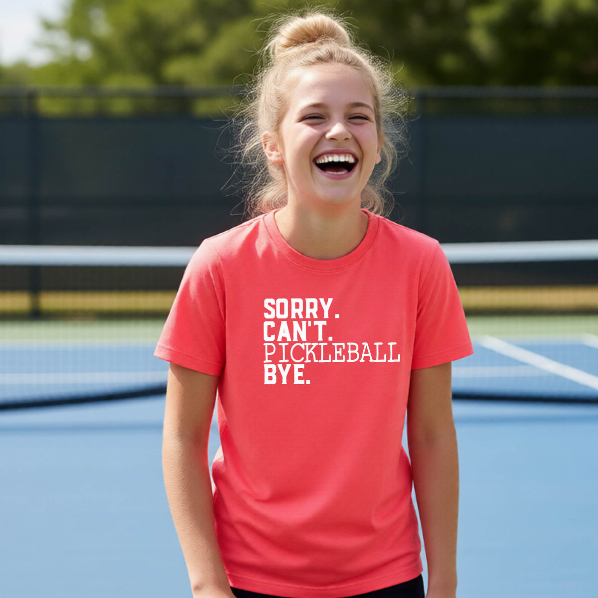 A young person on a pickleball court wearing a coral pink short-sleeve t-shirt with a white graphic of the words  “SORRY. CAN’T. PICKLEBALL BYE” each on a separate line, on the front of the shirt. 