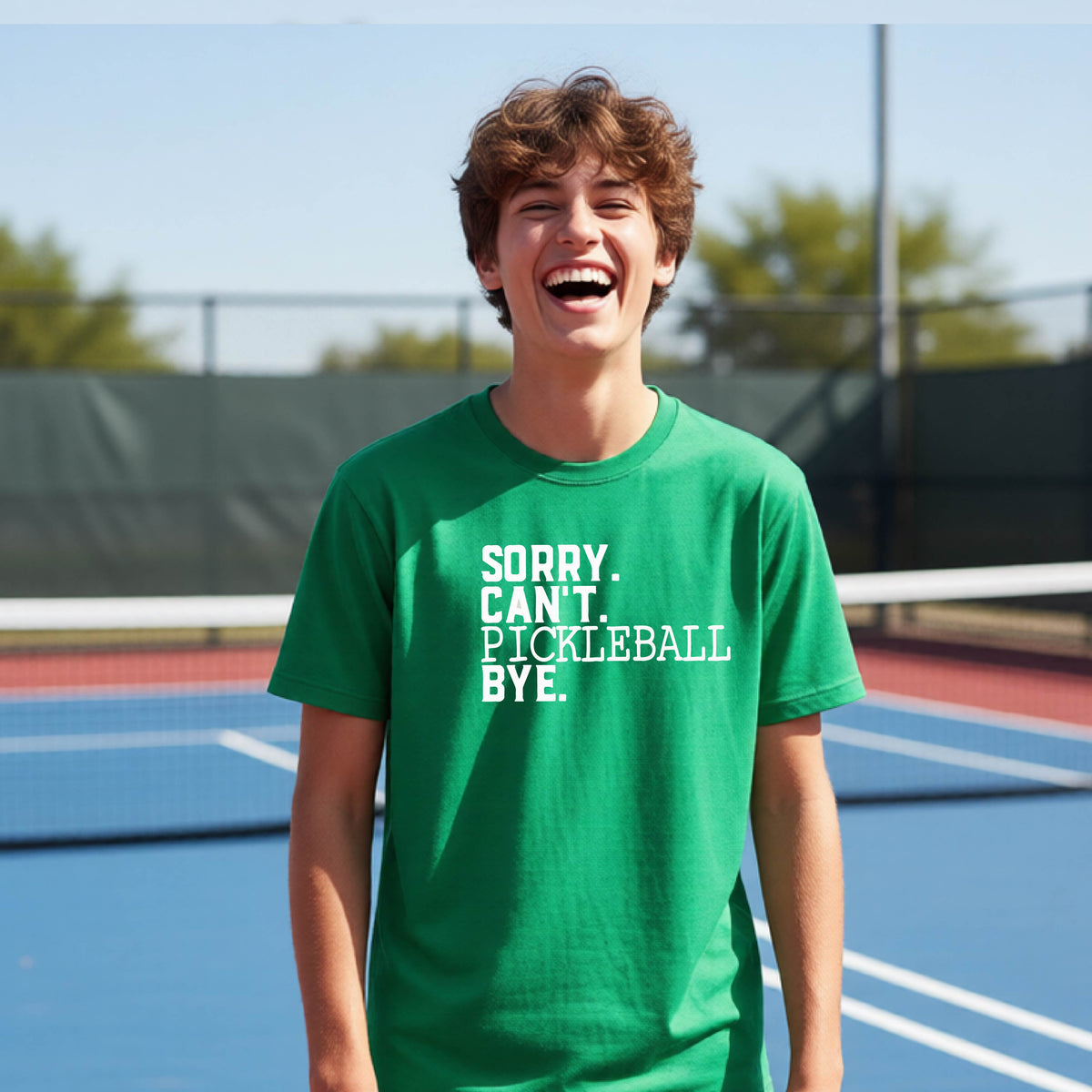A young person on a pickleball court wearing a green short-sleeve t-shirt with a white graphic of the words  “SORRY. CAN’T. PICKLEBALL BYE” each on a separate line, on the front of the shirt. 