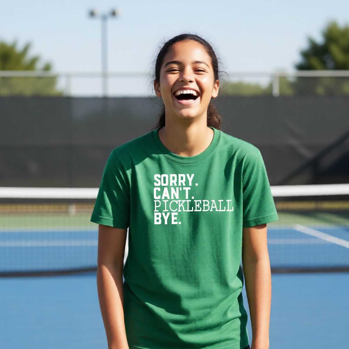 A young person on a pickleball court wearing a green short-sleeve t-shirt with a white graphic of the words  “SORRY. CAN’T. PICKLEBALL BYE” each on a separate line, on the front of the shirt. 