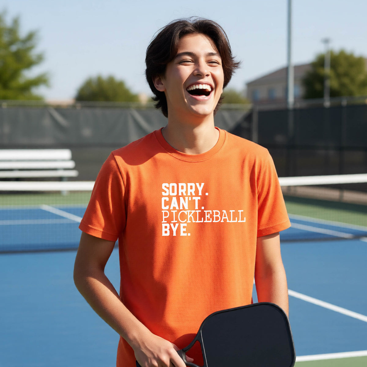 A young person on a pickleball court wearing an orange short-sleeve t-shirt with a white graphic of the words  “SORRY. CAN’T. PICKLEBALL BYE” each on a separate line, on the front of the shirt. 