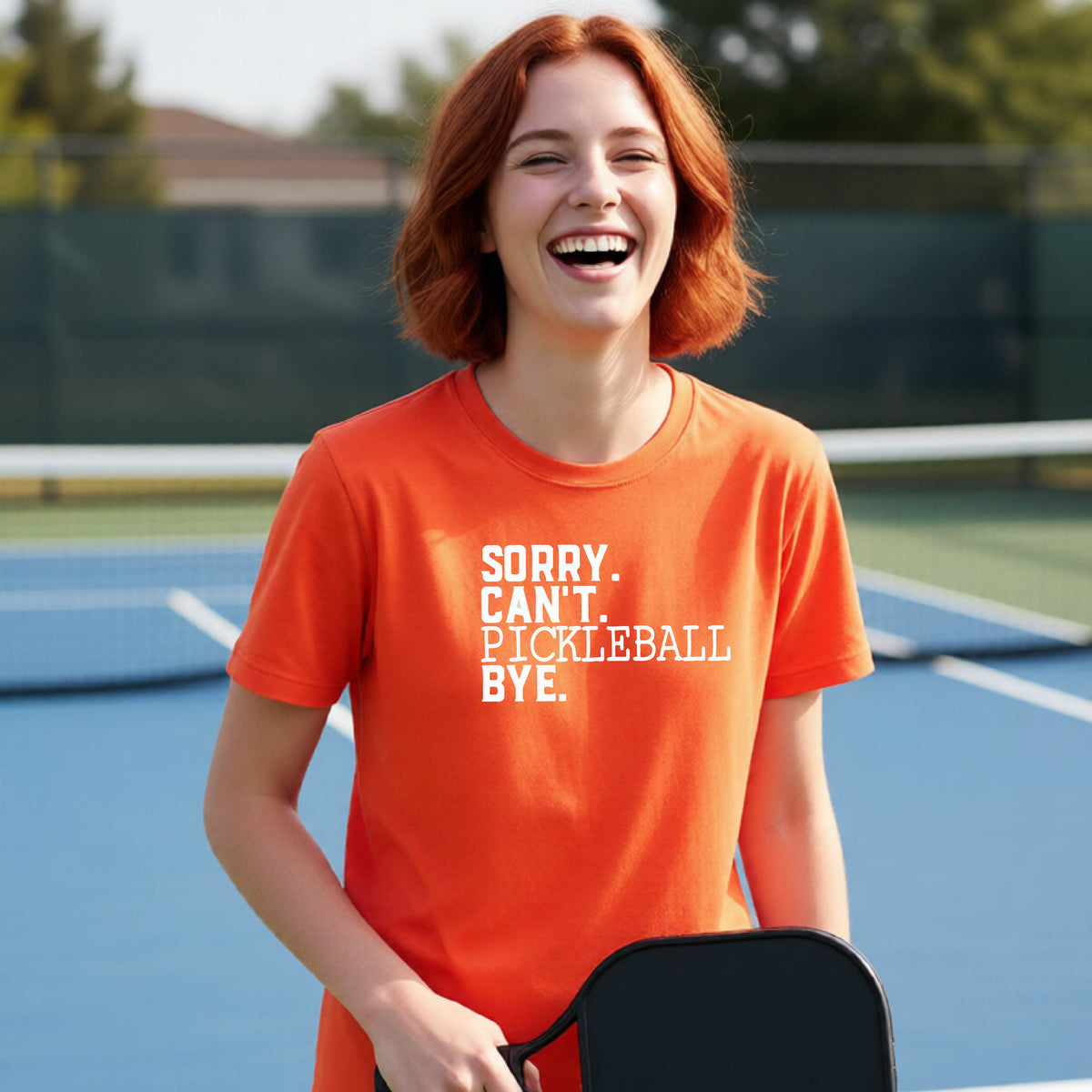 A young person on a pickleball court wearing an orange short-sleeve t-shirt with a white graphic of the words  “SORRY. CAN’T. PICKLEBALL BYE” each on a separate line, on the front of the shirt. 